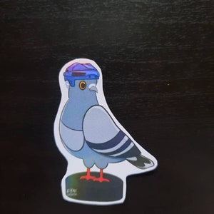 May include: A blue and white pigeon wearing a blue hockey helmet with a yellow visor. The pigeon is standing on a black and white base with the text "KMC CREATIONS" printed on it.