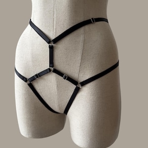 May include: A black harness made of thin elastic straps and silver-toned metal rings and sliders. The design features a geometric arrangement of interconnected bands that wrap around the waist and hips, creating a structured, cage-like appearance.