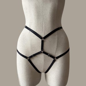 May include: A black elastic harness featuring thin adjustable straps and silver-toned metal rings. The design consists of a waist band connected to a central vertical strap and a lower V-shaped section, creating a geometric pattern across the torso.