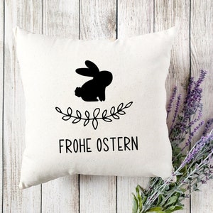 Peut inclure: Un oreiller blanc avec une silhouette noire de lapin et une couronne de feuilles. Le texte "Frohe Ostern" est imprim&eacute; sous le lapin.