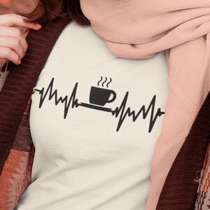 Puede incluir: Camiseta blanca con un gr&aacute;fico negro de una taza de caf&eacute; con vapor saliendo de ella y una l&iacute;nea de latido del coraz&oacute;n a cada lado de la taza. El texto "Me encanta el caf&eacute;" no es visible en la imagen.