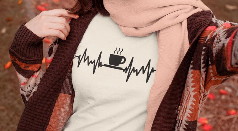 Puede incluir: Camiseta blanca con un gr&aacute;fico negro de una taza de caf&eacute; con vapor saliendo de ella y una l&iacute;nea de latido del coraz&oacute;n a cada lado de la taza. El texto "Me encanta el caf&eacute;" no es visible en la imagen.
