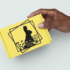 May include: A yellow box with a black silhouette of a rabbit sitting in a field of flowers. The rabbit is surrounded by a black square frame.