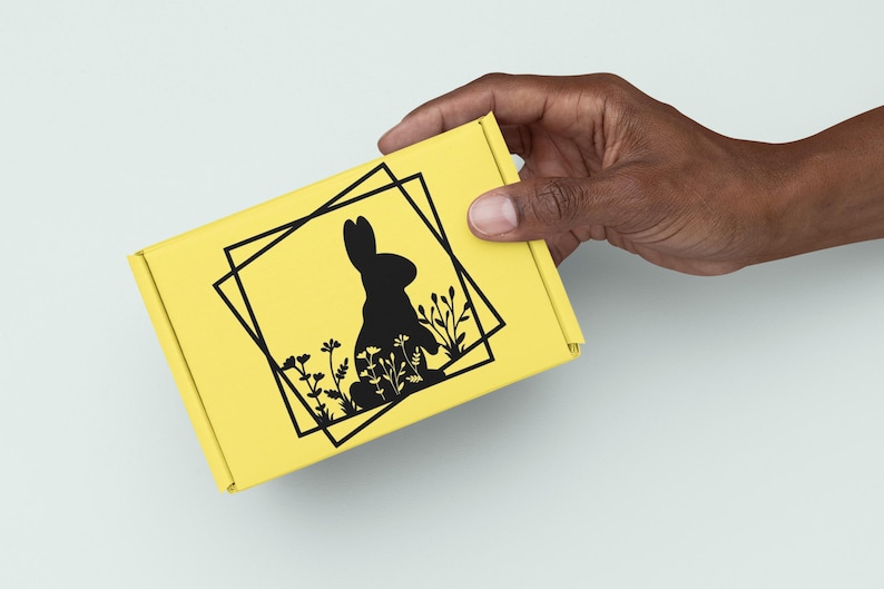 May include: A yellow box with a black silhouette of a rabbit sitting in a field of flowers. The rabbit is surrounded by a black square frame.