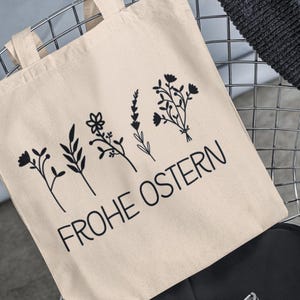 May include: A beige tote bag with black floral designs and the text "Frohe Ostern" printed on it.