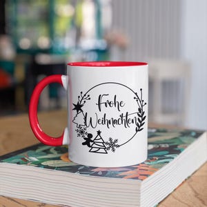 May include: White ceramic mug with a red rim. The mug has a black design of a Christmas tree, angel, snowflakes, and the text "Frohe Weihnachten" in a circle.