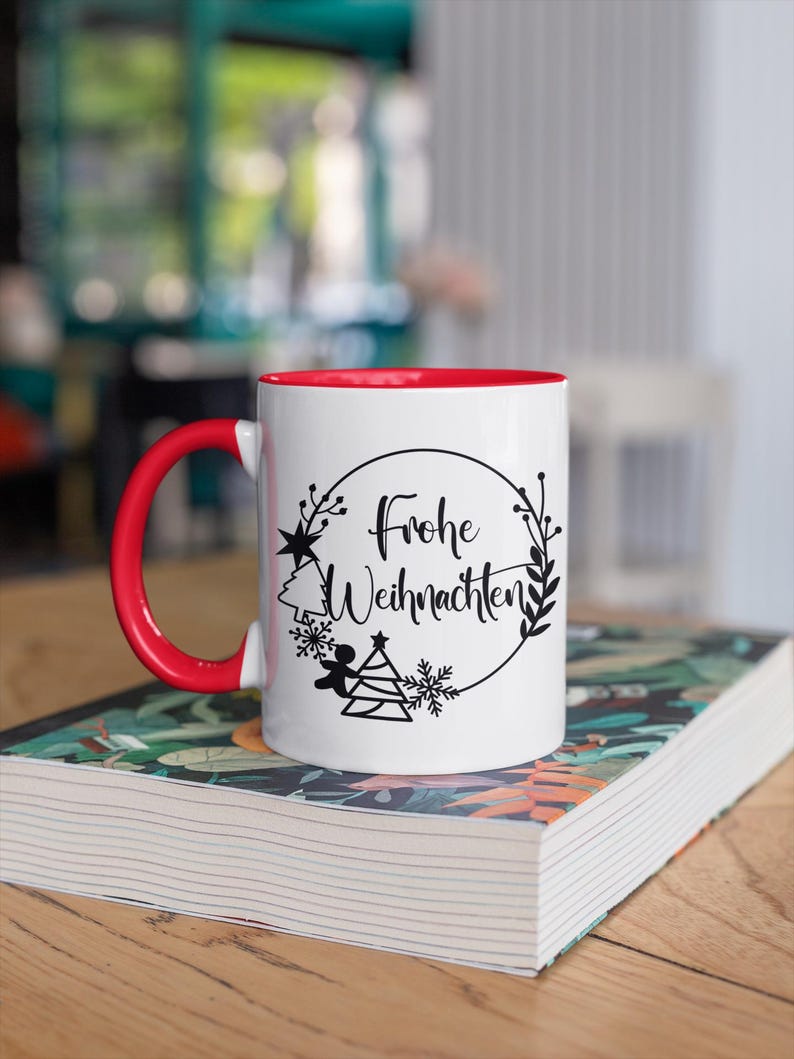 May include: White ceramic mug with a red rim. The mug has a black design of a Christmas tree, angel, snowflakes, and the text "Frohe Weihnachten" in a circle.