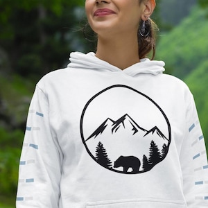 May include: A white hooded sweatshirt featuring a black circular graphic on the chest. The graphic depicts a mountain range above a forest with a bear silhouette. The sleeves have a repeating pattern of small light blue and grey rectangular shapes.