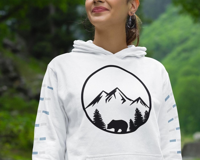 May include: A white hooded sweatshirt featuring a black circular graphic on the chest. The graphic depicts a mountain range above a forest with a bear silhouette. The sleeves have a repeating pattern of small light blue and grey rectangular shapes.
