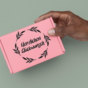 K&ouml;nnte beinhalten: Eine rosa Geschenkbox mit einem schwarzen floralen Kranz und dem Text "Herzlichen Gl&uuml;ckwunsch".
