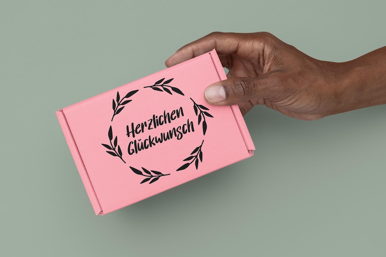 K&ouml;nnte beinhalten: Eine rosa Geschenkbox mit einem schwarzen floralen Kranz und dem Text "Herzlichen Gl&uuml;ckwunsch".