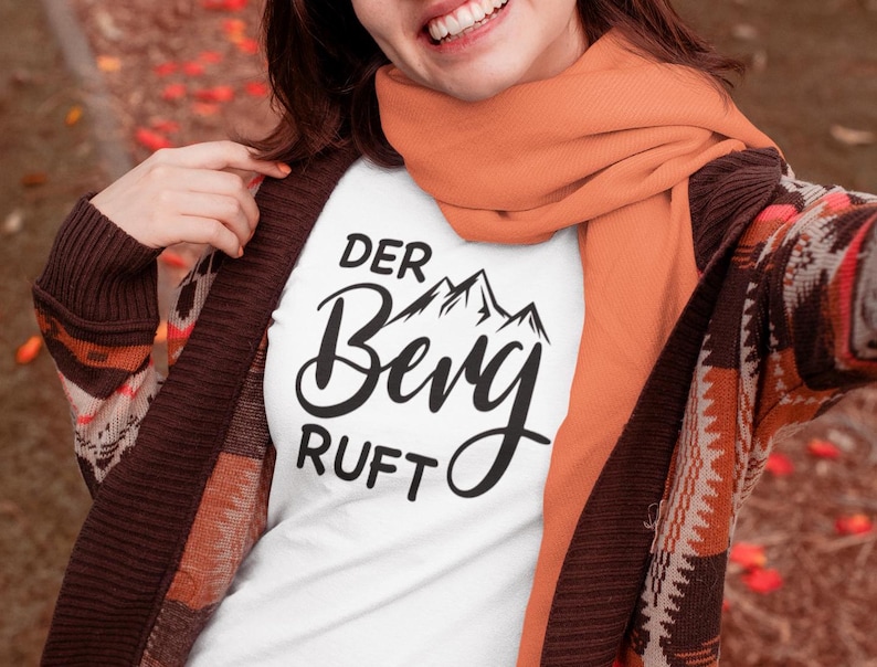May include: A white t-shirt featuring black text that reads "DER BERG RUFT" with a graphic of mountain peaks above the word "Berg". The shirt is worn under a brown patterned cardigan and an orange scarf, creating a layered look.