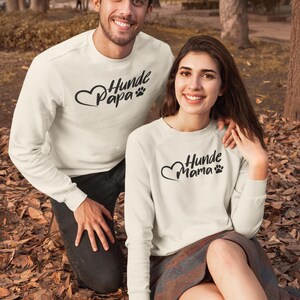 May include: Two people wearing matching cream-colored sweatshirts with the text "Hunde Papa" and "Hunde Mama" in black script, along with a heart and paw print design. They are outdoors in a park setting.