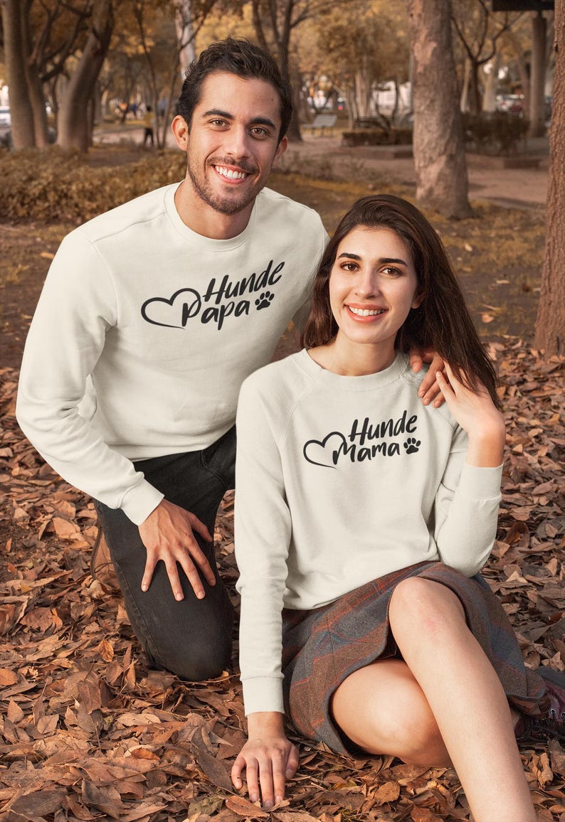May include: Two people wearing matching cream-colored sweatshirts with the text "Hunde Papa" and "Hunde Mama" in black script, along with a heart and paw print design. They are outdoors in a park setting.