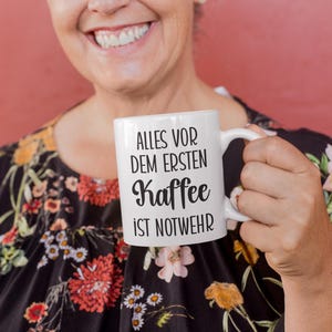以下が含まれることがあります： ドイツ語のテキスト「ALLES VOR DEM ERSTEN Kaffee IST NOTWEHR」が黒いスクリプトで書かれた白いセラミックコーヒーマグ。マグカップは、赤い背景に花柄のトップスを着た人が持っています。マグカップは標準サイズです。
