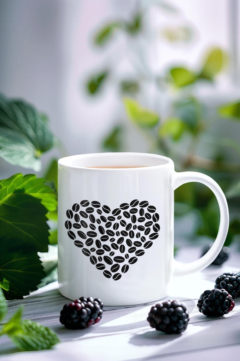 以下が含まれることがあります： 黒いコーヒー豆でできたハートのデザインが施された白いセラミックコーヒーマグ。 マグカップには茶色の液体が入っており、明るい色の表面に置かれ、周りにはブラックベリーが散らばっています。