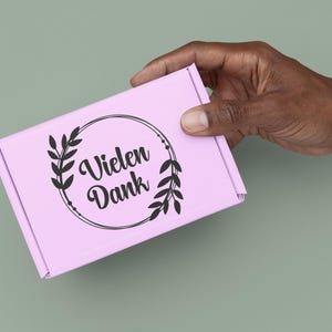 May include: A light pink rectangular cardboard shipping box featuring a black circular graphic design with two leafy branches on the sides and the German text "Vielen Dank" printed in a stylised script font in the centre of the circle.