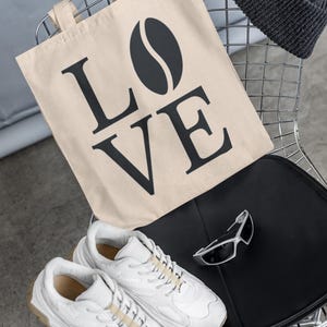 以下が含まれることがあります： 「LOVE」の文字とコーヒー豆のグラフィックが入ったベージュのトートバッグ、濃いグレーのビーニー、白いスニーカー、シルバーのサングラスが、黒い座面のある金属製の椅子に置かれています。