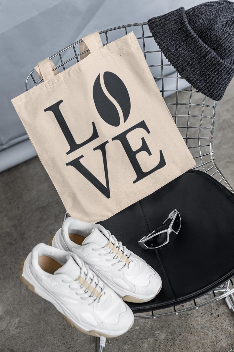 以下が含まれることがあります： 「LOVE」の文字とコーヒー豆のグラフィックが入ったベージュのトートバッグ、濃いグレーのビーニー、白いスニーカー、シルバーのサングラスが、黒い座面のある金属製の椅子に置かれています。
