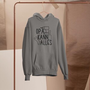 May include: A grey hoodie with a front pocket and a drawstring hood is hanging on a copper-coloured clothing rack. The hoodie features the text "OPA KANN ALLES" in black, with tool illustrations.