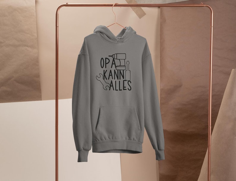 May include: A grey hoodie with a front pocket and a drawstring hood is hanging on a copper-coloured clothing rack. The hoodie features the text "OPA KANN ALLES" in black, with tool illustrations.