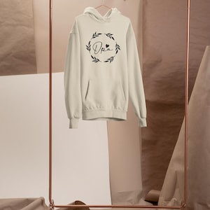 May include: Cream hoodie with a front pocket and hood, displayed on a copper clothes rail. The hoodie has the word "Opa" in a decorative font, surrounded by a wreath design. The background includes paper rolls and sheets.