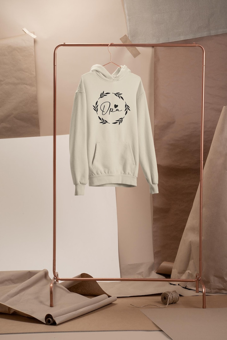 May include: Cream hoodie with a front pocket and hood, displayed on a copper clothes rail. The hoodie has the word "Opa" in a decorative font, surrounded by a wreath design. The background includes paper rolls and sheets.
