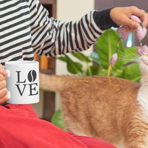 以下が含まれることがあります： 「LOVE」の文字とコーヒー豆のグラフィックが入った白いコーヒーマグ。白と黒のストライプのシャツを着た人がマグカップを持っています。オレンジと白の猫がピンクのおもちゃで遊んでいます。