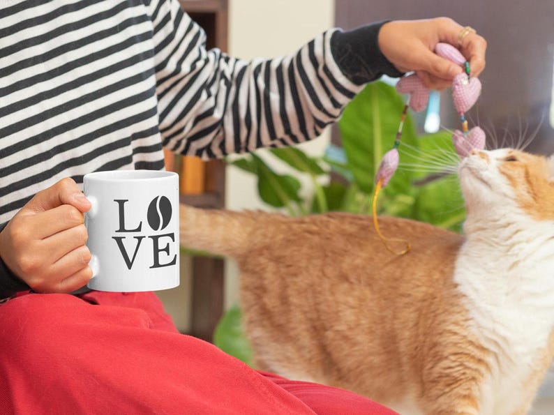 以下が含まれることがあります： 「LOVE」の文字とコーヒー豆のグラフィックが入った白いコーヒーマグ。白と黒のストライプのシャツを着た人がマグカップを持っています。オレンジと白の猫がピンクのおもちゃで遊んでいます。