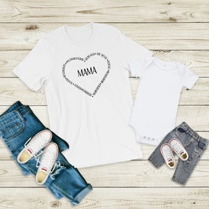 Peut inclure: T-shirt blanc avec un motif en forme de c&oelig;ur qui dit "MAMA" &agrave; l'int&eacute;rieur et des mots autour du c&oelig;ur. Un body blanc pour b&eacute;b&eacute; avec un motif assorti. Les deux t-shirts sont sur un fond en bois avec un jean bleu et des baskets blanches.