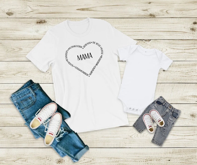 Peut inclure: T-shirt blanc avec un motif en forme de c&oelig;ur qui dit "MAMA" &agrave; l'int&eacute;rieur et des mots autour du c&oelig;ur. Un body blanc pour b&eacute;b&eacute; avec un motif assorti. Les deux t-shirts sont sur un fond en bois avec un jean bleu et des baskets blanches.