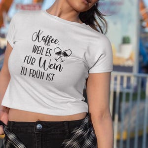 以下が含まれることがあります： ドイツ語のテキスト「Kaffee, weil es f&uuml;r Wein zu fr&uuml;h ist」とワイングラスのグラフィックが入った白いクロップトップTシャツ。カジュアルなスタイルのシャツです。