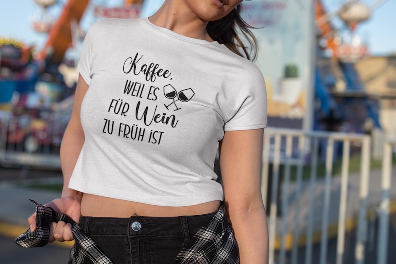 以下が含まれることがあります： ドイツ語のテキスト「Kaffee, weil es f&uuml;r Wein zu fr&uuml;h ist」とワイングラスのグラフィックが入った白いクロップトップTシャツ。カジュアルなスタイルのシャツです。