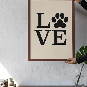 May include: A framed print with the word "LOVE" in black capital letters and a black paw print replacing the "O". The frame is brown, and the background is a light beige color. The print is held up by a person.