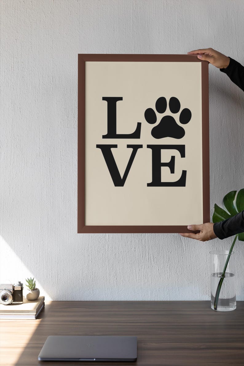 May include: A framed print with the word "LOVE" in black capital letters and a black paw print replacing the "O". The frame is brown, and the background is a light beige color. The print is held up by a person.