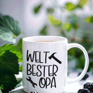 May include: White ceramic mug with the text "WELT BESTER OPA" in black, featuring a hammer and wrench graphic. The mug contains a brown liquid and is set amongst blackberries and green leaves.
