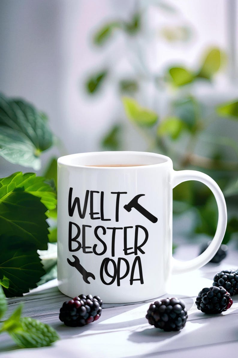 May include: White ceramic mug with the text "WELT BESTER OPA" in black, featuring a hammer and wrench graphic. The mug contains a brown liquid and is set amongst blackberries and green leaves.