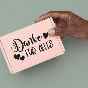 May include: A light pink rectangular cardboard shipping box featuring the black printed text "Danke F&Uuml;R ALLES" and three black heart icons. The box has a fold-over lid design and is constructed from a smooth, matte-finish paper material.
