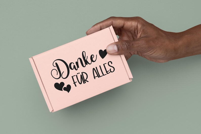 May include: A light pink rectangular cardboard shipping box featuring the black printed text "Danke F&Uuml;R ALLES" and three black heart icons. The box has a fold-over lid design and is constructed from a smooth, matte-finish paper material.