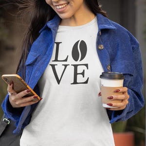 以下が含まれることがあります： 「LOVE」の文字が黒いブロック体で書かれ、「O」の代わりにコーヒー豆のグラフィックが描かれた白いTシャツ。青いコーデュロイのジャケットを着て、スマートフォンとコーヒーカップを持っています。