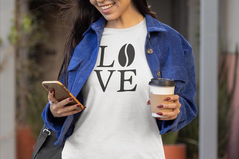 以下が含まれることがあります： 「LOVE」の文字が黒いブロック体で書かれ、「O」の代わりにコーヒー豆のグラフィックが描かれた白いTシャツ。青いコーデュロイのジャケットを着て、スマートフォンとコーヒーカップを持っています。