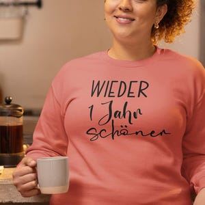 K&ouml;nnte beinhalten: Ein korallenfarbenes Sweatshirt mit dem Aufdruck "WIEDER 1 Jahr sch&ouml;ner" in Schwarz.
