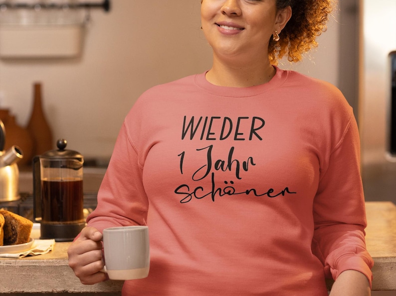 K&ouml;nnte beinhalten: Ein korallenfarbenes Sweatshirt mit dem Aufdruck "WIEDER 1 Jahr sch&ouml;ner" in Schwarz.