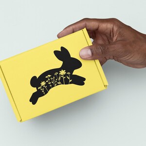 May include: A yellow box with a black silhouette of a rabbit with flowers inside. The box is being held by a hand.
