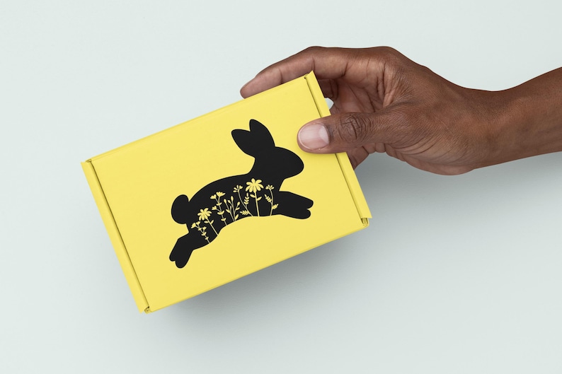 May include: A yellow box with a black silhouette of a rabbit with flowers inside. The box is being held by a hand.