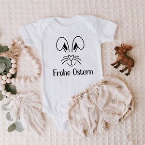 Peut inclure: Combinaison blanche pour b&eacute;b&eacute; avec un visage de lapin noir et le texte "Frohe Ostern" en noir.