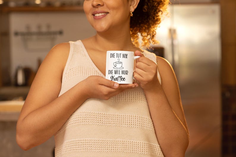 以下が含まれることがあります： 黒い文字とコーヒーカップのグラフィックが入った白いセラミックコーヒーマグ。マグには「DIE TUT NIX, DIE WILL NUR KAFFEE」と書かれています。マグは、クリーム色のタンクトップを着た人が持っています。