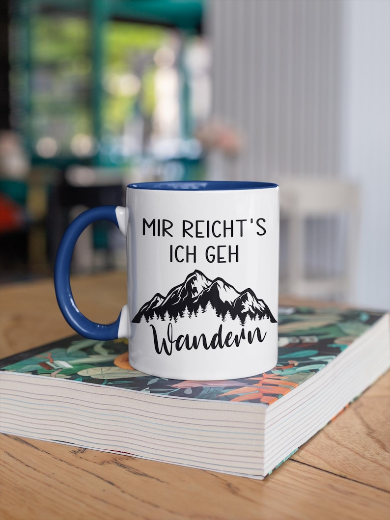 May include: A white ceramic mug with a dark blue handle and interior. The front features black text that reads "MIR REICHT'S ICH GEH" above a black graphic illustration of a mountain range with pine trees and the word "Wandern" below it.