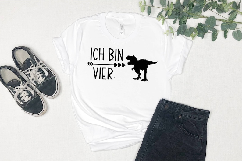 May include: White t-shirt with black text that reads "Ich bin vier" and a black silhouette of a dinosaur. The dinosaur is facing left and has its mouth open. An arrow points to the dinosaur's mouth.