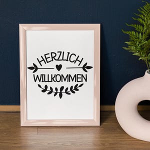 Op de afbeelding: Een roze ingelijste print met de tekst "HERZLICH WILLKOMMEN" in zwarte letters met een klein hartje en een simpel pijlontwerp. De print is omringd door een krans van bladeren.